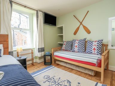 A bedroom with a wooden daybed, striped cushions, a wooden floor, a window with curtains, a wall-mounted TV, and two crossed wooden paddles on the wall at Gadlys House - Waters Edge Beaumaris