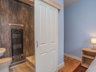 A bathroom with a toilet sink and shower visible through a partially open white sliding door next to a bedroom with a wooden nightstand and lamp at Gadlys House - Waters Edge in Beaumaris