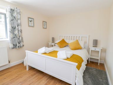 A bedroom with a white bed frame yellow pillows and a blanket two white bedside tables with lamps floral curtains and wooden floor at Puddleducks in Sherborne