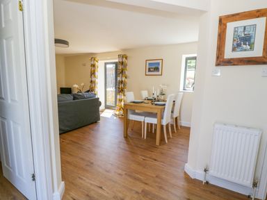 A dining area with a wooden table and white chairs next to a living room with a grey sofa and patterned curtains at Puddleducks in Sherborne