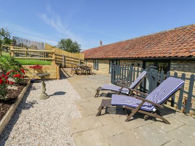 An outdoor patio with two lounge chairs, a dining table, a birdbath, flowers, and a stone building at Puddleducks in Sherborne