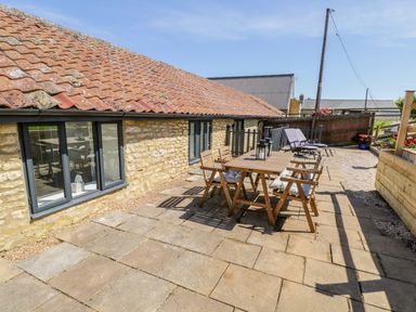 An outdoor patio area with a wooden table and four chairs next to a stone building with a tiled roof and windows at Puddleducks Sherborne