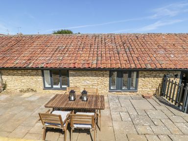 An outdoor patio with a wooden table and four chairs in front of a stone building with red tiled roof at Puddleducks in Sherborne