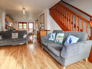 A living room with two grey sofas with blue and patterned cushions a wooden staircase and a dining area in the background at Sunflower Cottage in St Issey