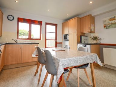 A kitchen with wooden cabinets a dining table with chairs and a door leading outside at Sunflower Cottage in St Issey
