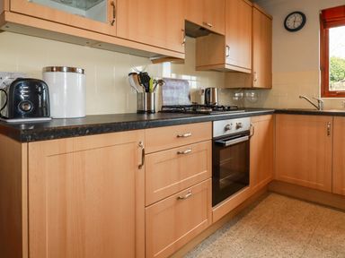 A kitchen with wooden cabinets a black countertop a toaster a bread bin a stove an oven and a sink by a window at Sunflower Cottage in St Issey