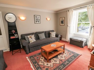 A living room with a grey sofa, wooden coffee table, patterned rug, tall black clock shelf, and a window with white curtains at Simonside Apartment in Rothbury