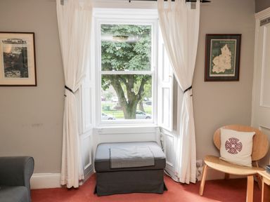 A sitting area with a window between white curtains a gray ottoman and a wooden chair with a cushion at Simonside Apartment in Rothbury