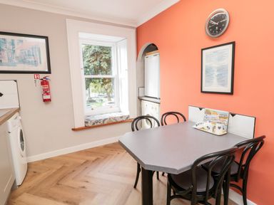 A kitchen dining area with a table and four chairs a window with a cushion washing machines and an orange wall with a clock and framed picture at Simonside Apartment in Rothbury
