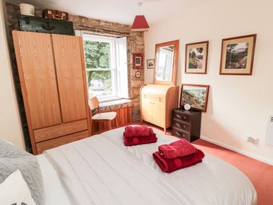 A bedroom with a bed with red towels on it a wooden wardrobe a chair by a window a wooden dresser with a mirror a small dark chest of drawers and framed pictures on the wall at Simonside Apartment in Rothbury