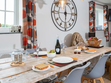 A dining table set with plates glasses a bottle and food with a large wall clock and patterned curtains at Bear Cottage in Chacewater near Truro