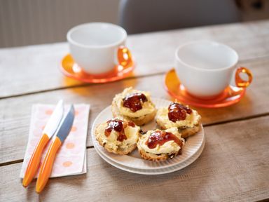 A wooden table with two white cups on orange saucers four scones with cream and jam on a white plate two knives with orange handles on a napkin at Bear Cottage in Chacewater near Truro