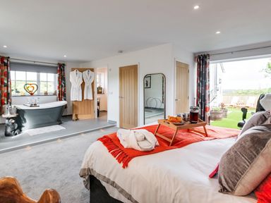 A bedroom with a bed and breakfast tray with bathrobes hanging on a door and a bathtub near a window at Bear Cottage in Chacewater near Truro