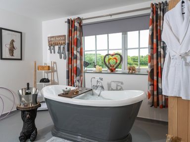 A bathroom with a gray bathtub filled with bubbles and a wooden tray with two glasses a window with curtains and decorative items on the sill a white bathrobe hanging and a bear-shaped stand holding a champagne bucket at Bear Cottage in Chacewater near Truro