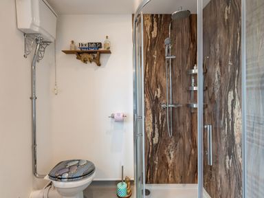 A bathroom with a toilet with a decorative lid and a shower with a glass door and wood-patterned walls at Bear Cottage in Chacewater near Truro