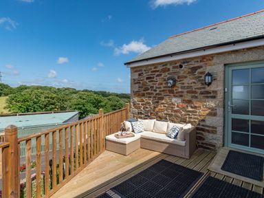 An outdoor wooden deck with a wicker sofa and cushions next to a stone wall and glass door at Bear Cottage in Chacewater near Truro