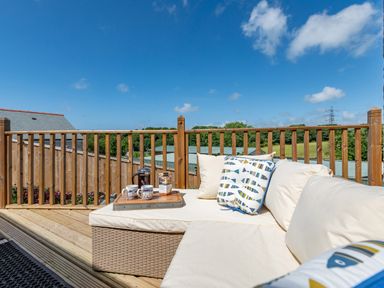 An outdoor wooden deck with a cushioned wicker sofa and a tray with mugs and coffee at Bear Cottage in Chacewater near Truro