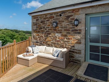 An outdoor seating area with a cushioned corner sofa on a wooden deck next to a stone wall with a door at Bear Cottage in Chacewater near Truro