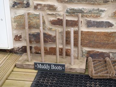 A wooden muddy boots rack with wooden pegs and a sign reading Muddy Boots on a decked floor by a stone wall