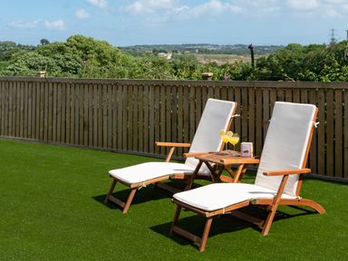 Two wooden lounge chairs with cushions and a small table with drinks on artificial grass with a wooden fence and trees at Bear Cottage in Chacewater near Truro