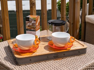 Two white cups with orange saucers on a wooden tray with a box of Cornish clotted cream and a glass pot on a wicker table on a wooden balcony at Bear Cottage in Chacewater near Truro