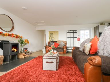 A living room with a red rug leather sofas a wood-burning stove and flowers on a table at Bear Cottage in Chacewater near Truro