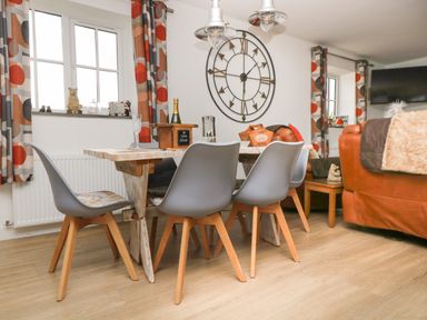 A dining area with a wooden table, gray chairs, a large wall clock, and patterned curtains at Bear Cottage in Chacewater near Truro