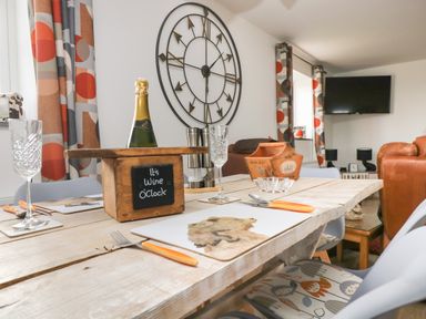 A dining area with a wooden table set with placemats glasses cutlery and a bottle under a sign saying Its Wine OClock wall clock and curtains with geometric patterns at Bear Cottage in Chacewater near Truro