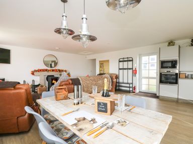 An open living and dining area with a wooden dining table set with placemats and glasses brown leather sofas a fireplace decorated with autumn leaves and a kitchen with built in oven and microwave at Bear Cottage in Chacewater near Truro