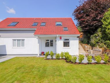A house with a red tiled roof white walls windows and a green lawn with plants and a bench outside at The Crest in Helston