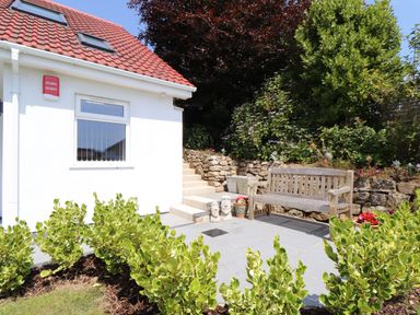A garden area with plants a wooden bench stone steps and a white house with a red tiled roof at The Crest in Helston