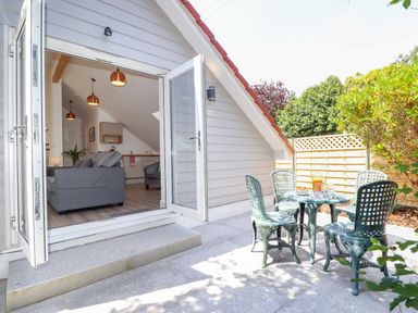 An open door leading to a living area with a sofa and pendant lights and an outdoor patio with green chairs and a table at The Crest in Helston