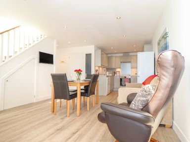 An open-plan room with a dining table and four chairs a kitchen area and two armchairs at The Crest in Helston