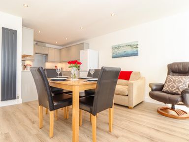 An open dining and living area with a wooden dining table set for four a beige sofa with a red throw and a brown armchair at The Crest in Helston