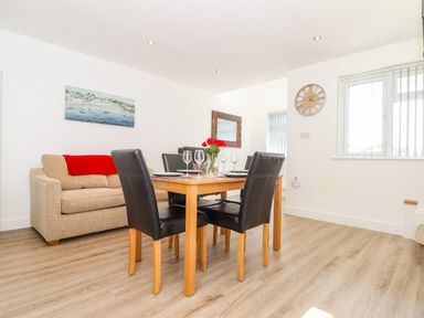 A dining area with a wooden table, four black chairs, a beige sofa with a red blanket, a wall clock, a mirror, and a window at The Crest in Helston