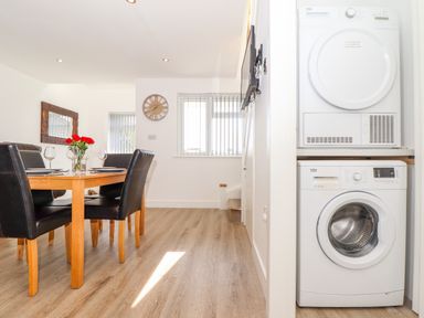 A dining area with a wooden table and black chairs next to stacked washer and dryer at The Crest in Helston
