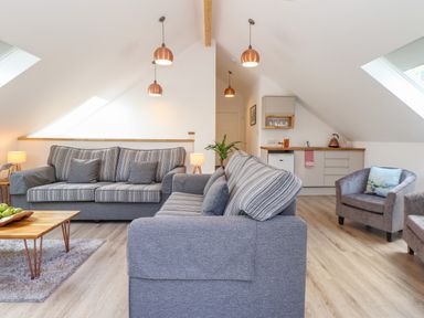 A living room with two grey striped sofas, two armchairs, a wooden coffee table with green apples, and a kitchenette at The Crest in Helston