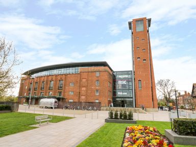 A brick building with a tall tower and a glass entrance with a welcome sign in a landscaped area with flowers and a bench at Booster's Lodge in Stratford-upon-Avon