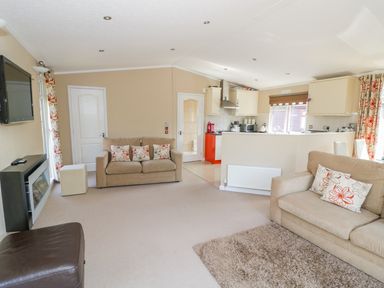 A living room with beige sofas and patterned cushions a wall-mounted TV and an open kitchen at Booster's Lodge in Stratford-upon-Avon