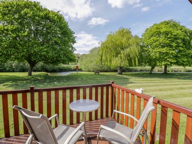 A wooden deck with two chairs and a small round table overlooking a grassy area with trees and a river at Boosters Lodge in Stratford-upon-Avon