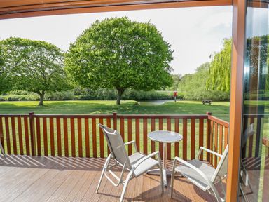 A wooden deck with two chairs and a small round table overlooking a grassy area with trees and a river at Booster's Lodge in Stratford-upon-Avon