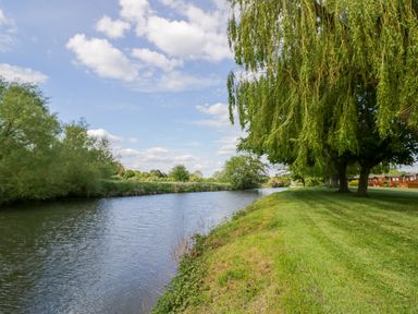 A river with grassy banks and trees under a partly cloudy sky at Boosters Lodge in Stratford-upon-Avon