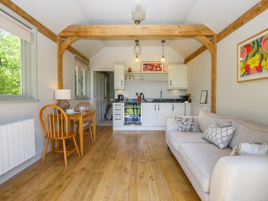A small living area with a wooden dining table and two chairs a beige sofa kitchen cabinets and appliances and wooden ceiling beams at The Lodge at Mortons in Hankerton