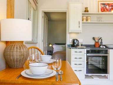 A dining table set with bowls, plates, utensils, and glasses next to a wicker lamp in a kitchen with white cabinets and an oven at The Lodge at Mortons in Hankerton