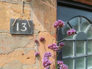 A house number 13 sign on a stone wall next to a window with textured glass and purple flowers in front at Abbey Cottage Montacute