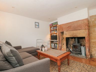 A living room with a gray sofa a wooden coffee table and a stone fireplace with a wood stove at Abbey Cottage Montacute