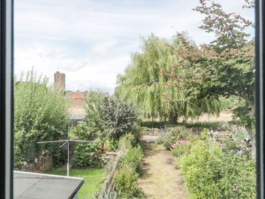 A garden with trees and shrubs and a path leading to a stone wall with a church tower in the background at Abbey Cottage Montacute