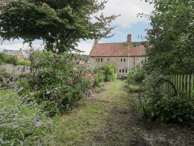 A garden with flowers and trees leading to a stone house with a tiled roof at Abbey Cottage Montacute