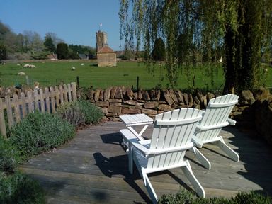 Two white wooden chairs and a small table on a wooden deck overlooking a field with sheep and a stone building at Abbey Cottage Montacute
