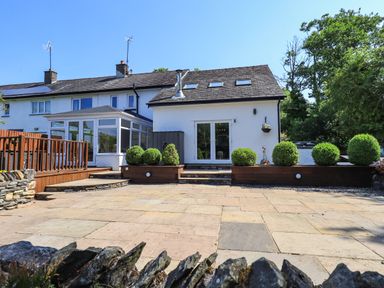 A backyard with paved patio area shrubs and a white house with glass doors and windows at Ellerbeck Bridge in Underbarrow near Kendal